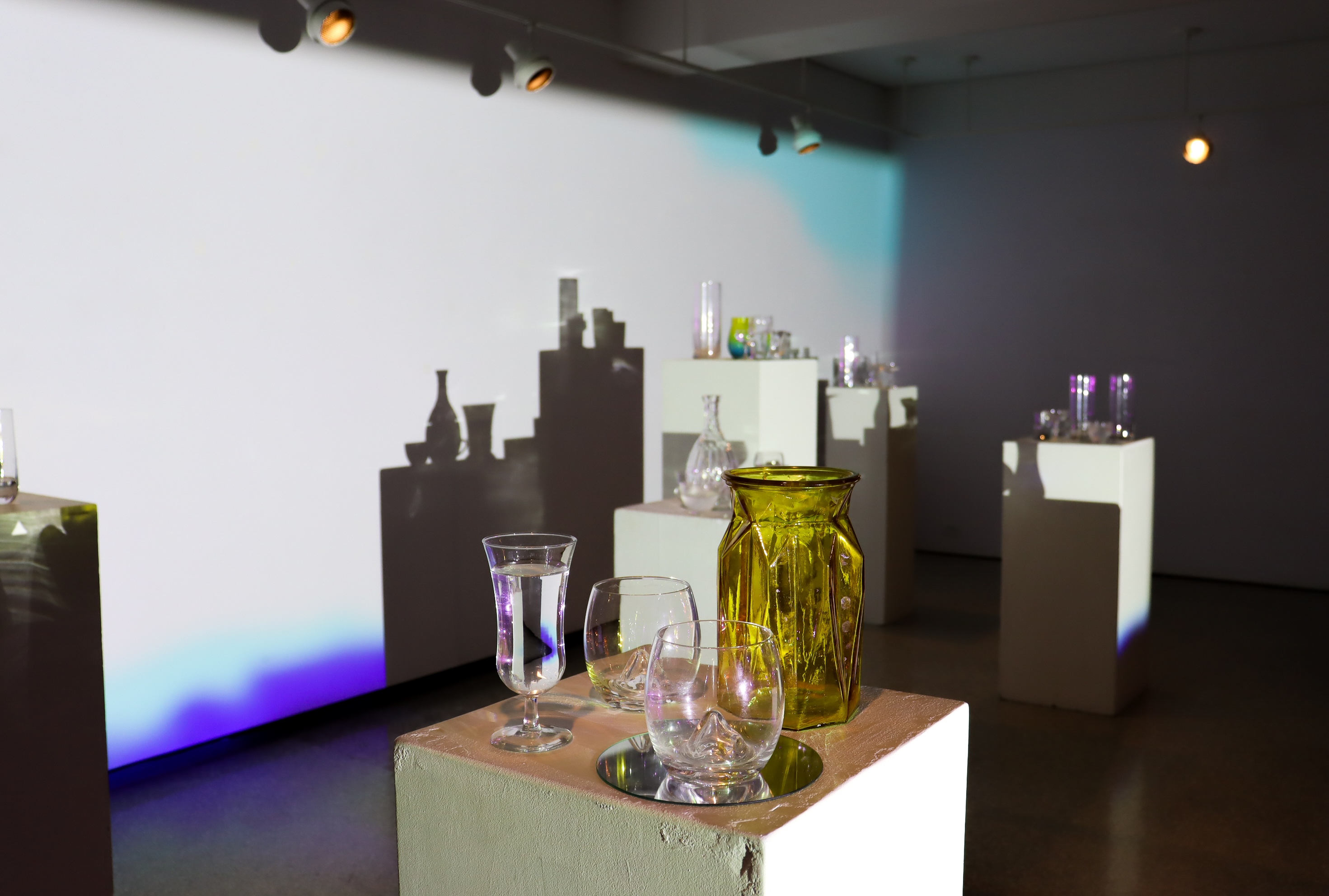 Close view of glassware and an amber vase on a plinth as sharp shadows and cool-toned projection stretch across the gallery wall.