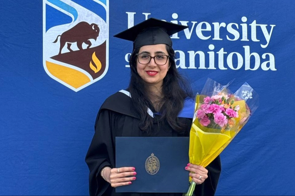A graduate in cap and gown holds her diploma and a bouquet of pink roses in yellow tissue