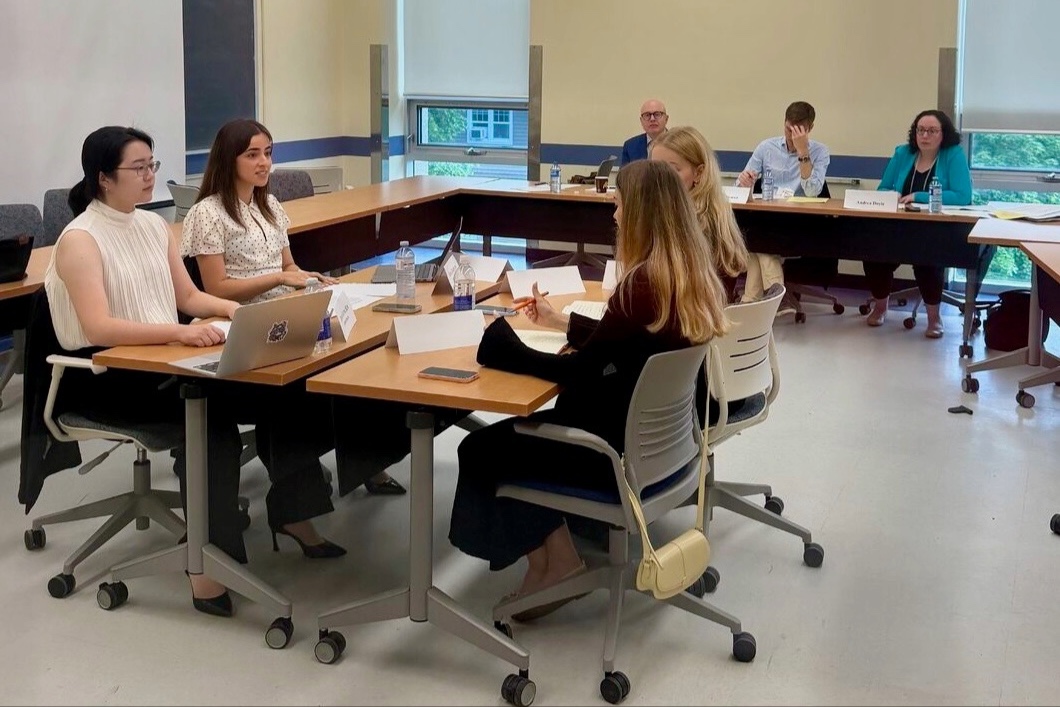 In a classroom, four students face each other at a table while a panel of three judges watch and listen