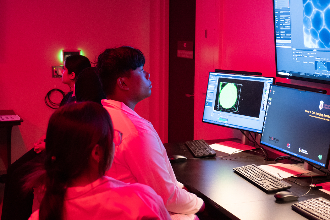 A student looks at computer screens. The room is lit by red lights.