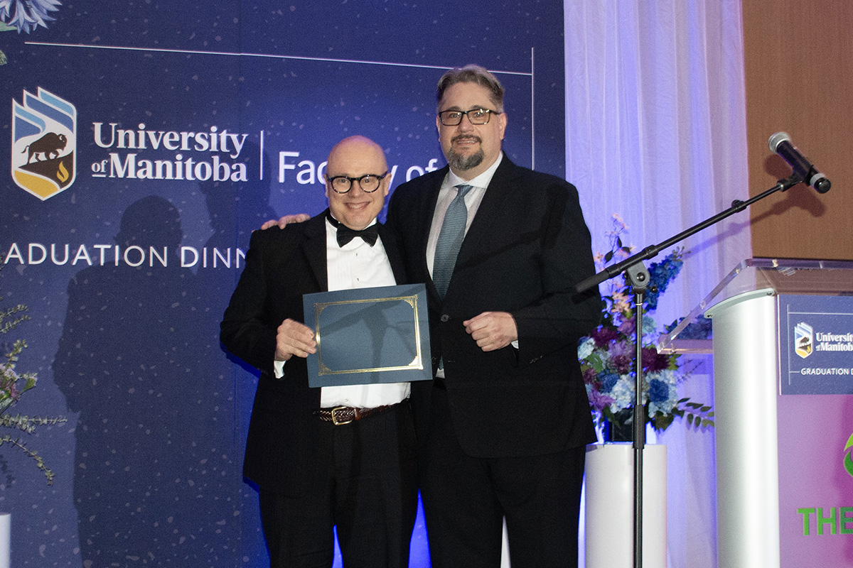 two men in suits smile on a stage one presenting an award certificate to the other