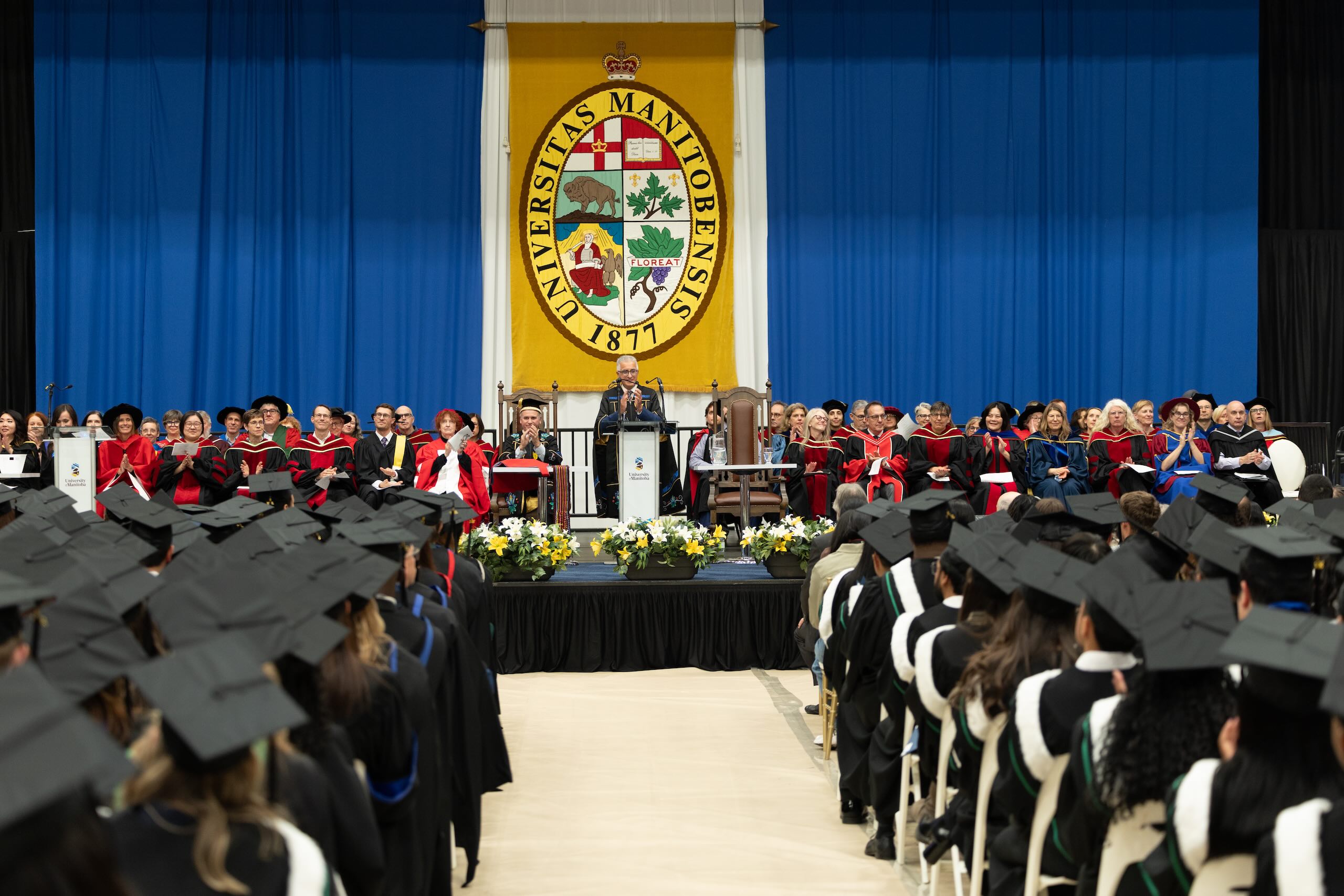 Convocation ceremony seen from back of room, with rows of students seated facing colourful stage with podium.