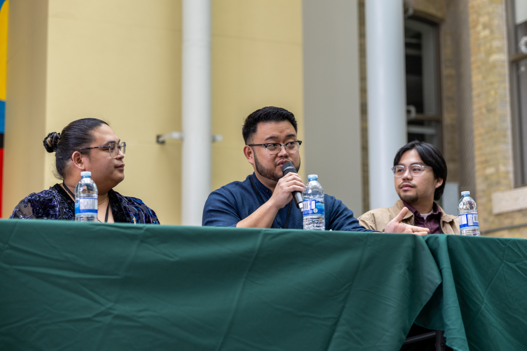 Three people sit behind a table. One person speaks into a microphone. 