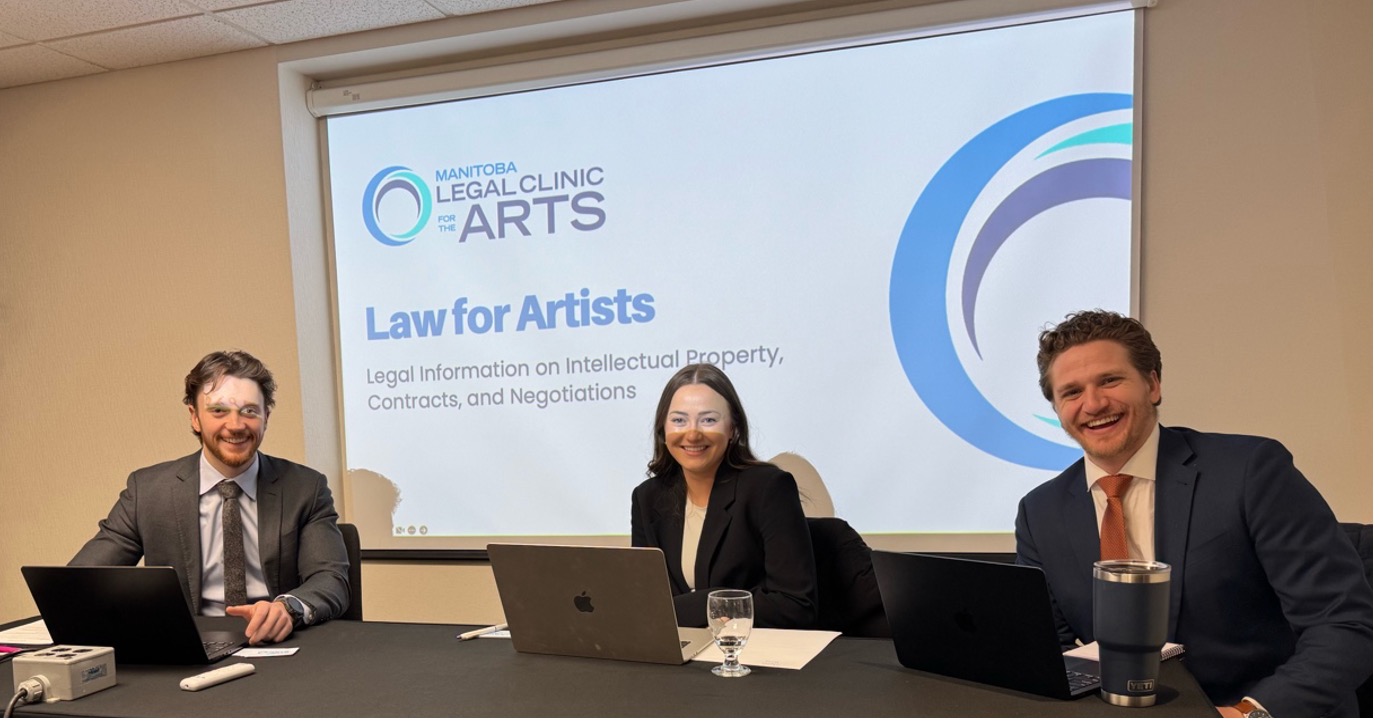 Three law students in business suits sit in front of laptop computers in front of a screen that says Manitoba Legal Clinic for the Arts Law for Artists