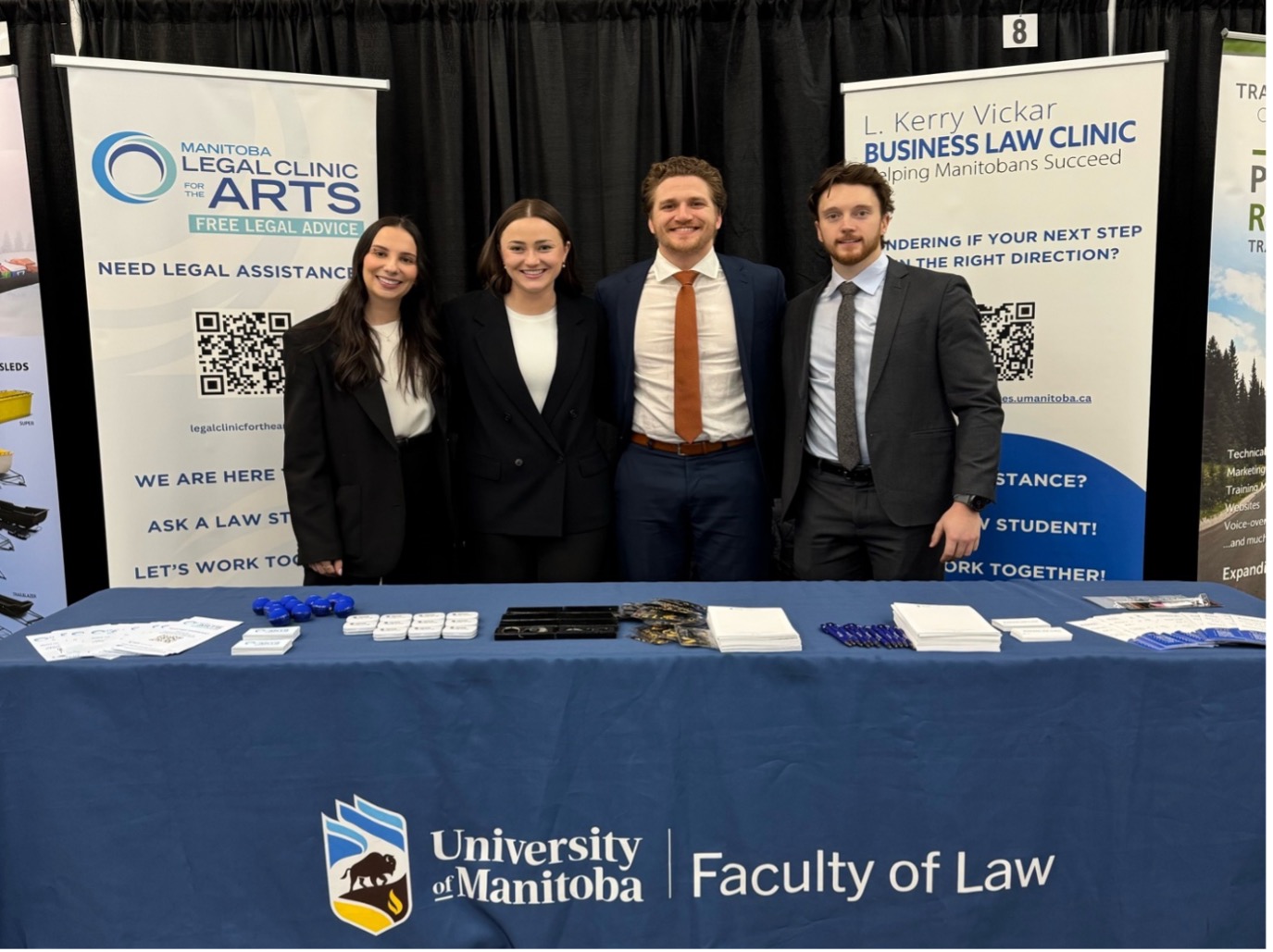 law students in suits and ties stand at an info booth between two roll up banners for the Kerry Vickar Business Law Clinic and the Arts Clinic