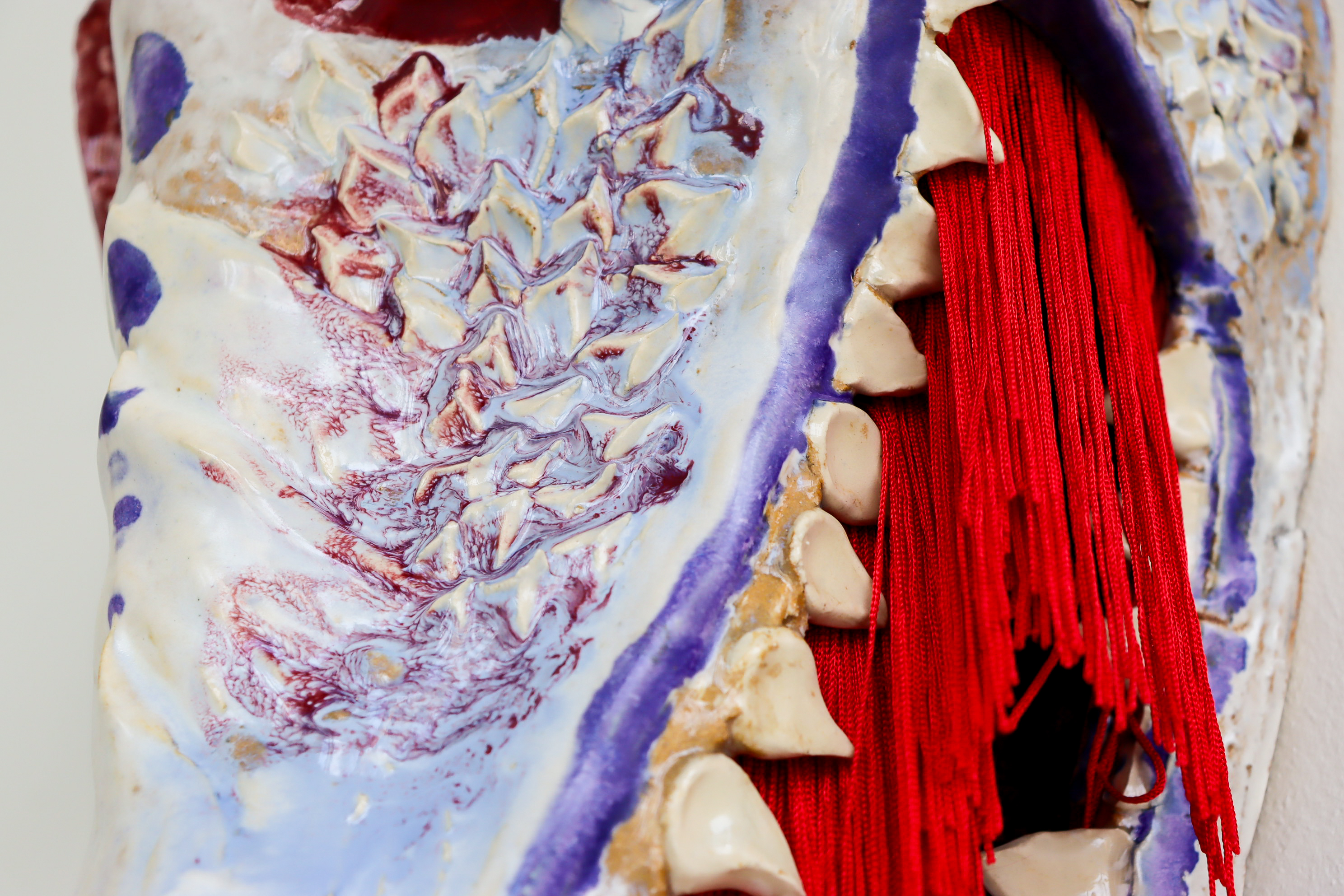 Close-up of a textured ceramic surface with raised forms and red thread elements woven through openings.