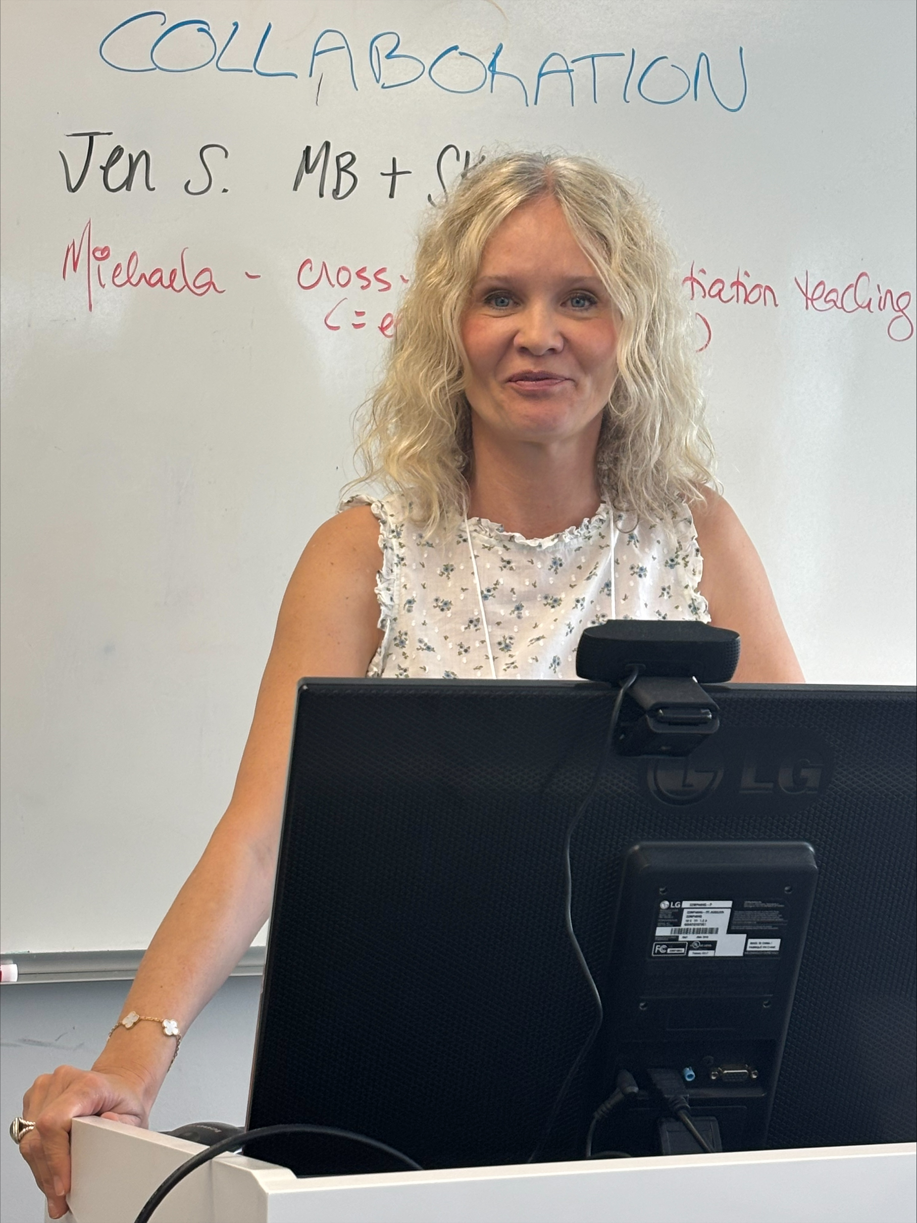 a woman in a white shirt gives a presentation in front of a white board from behind a computer monitor
