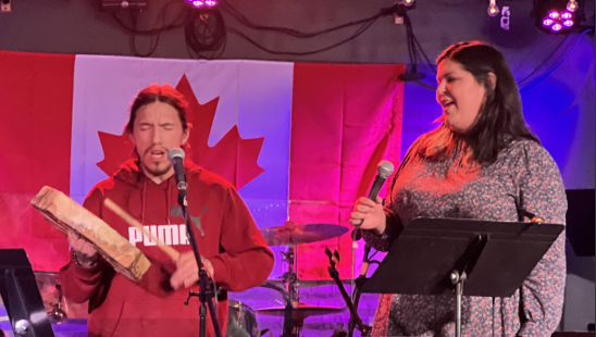 a man sings into a microphone while striking a flat handheld drum with a wood mallet and a woman sings into a microphone next to him