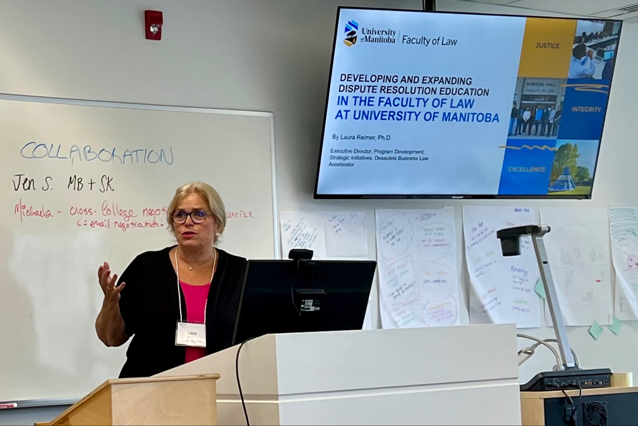 a woman in pink shirt and black sweater gives a presentation in front of a white board standing behind a computer screen.