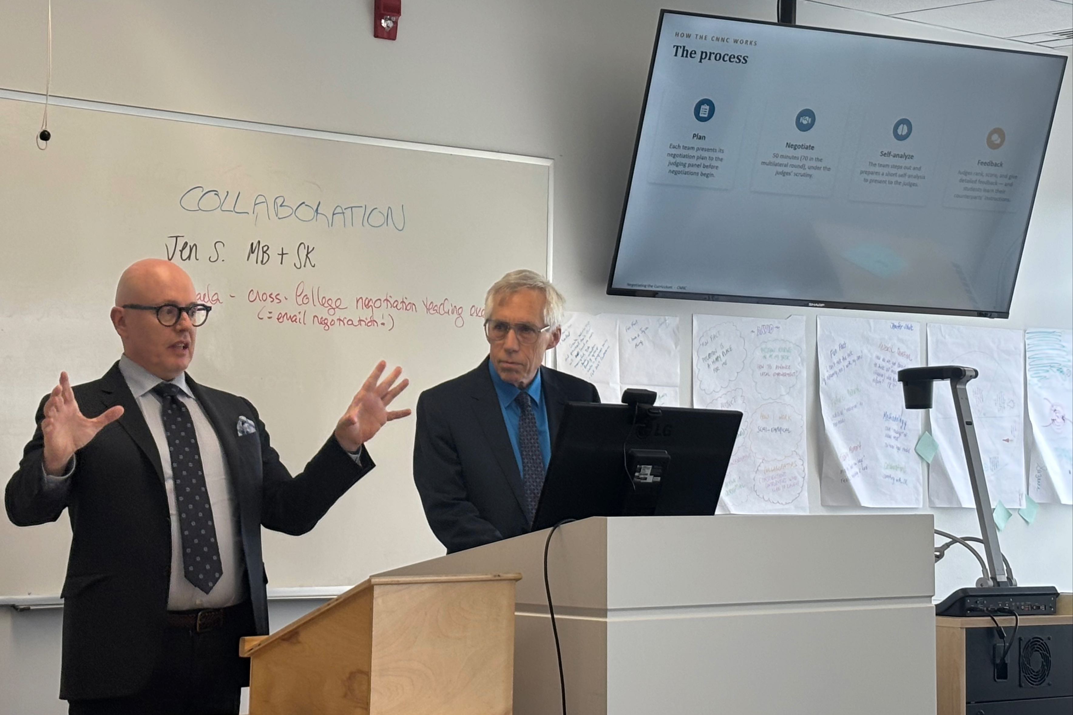 two men in suits give a presentation in front of a whiteboard behind a podium with computer screen