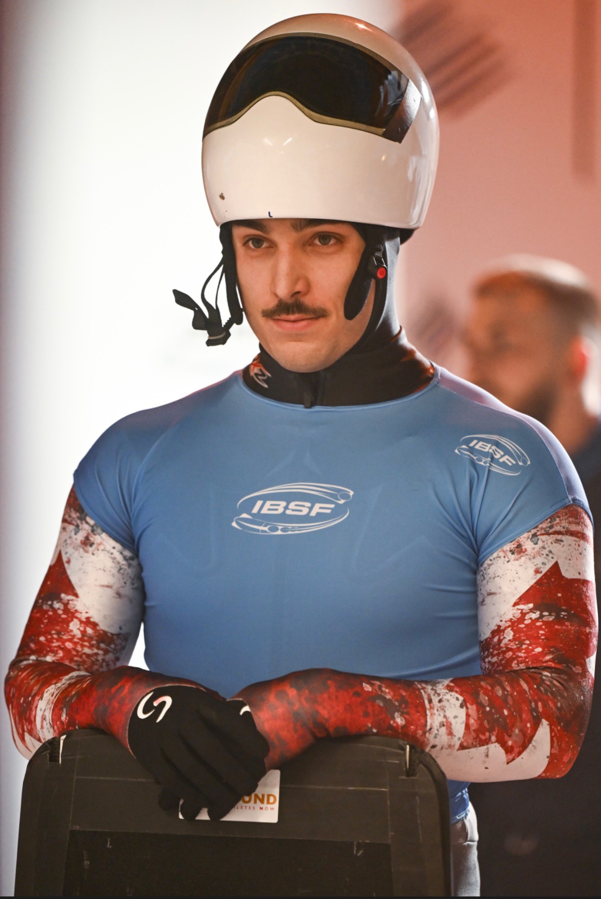 A man stands at the top of a skeleton track with his helment up, resting his hands on this sled.