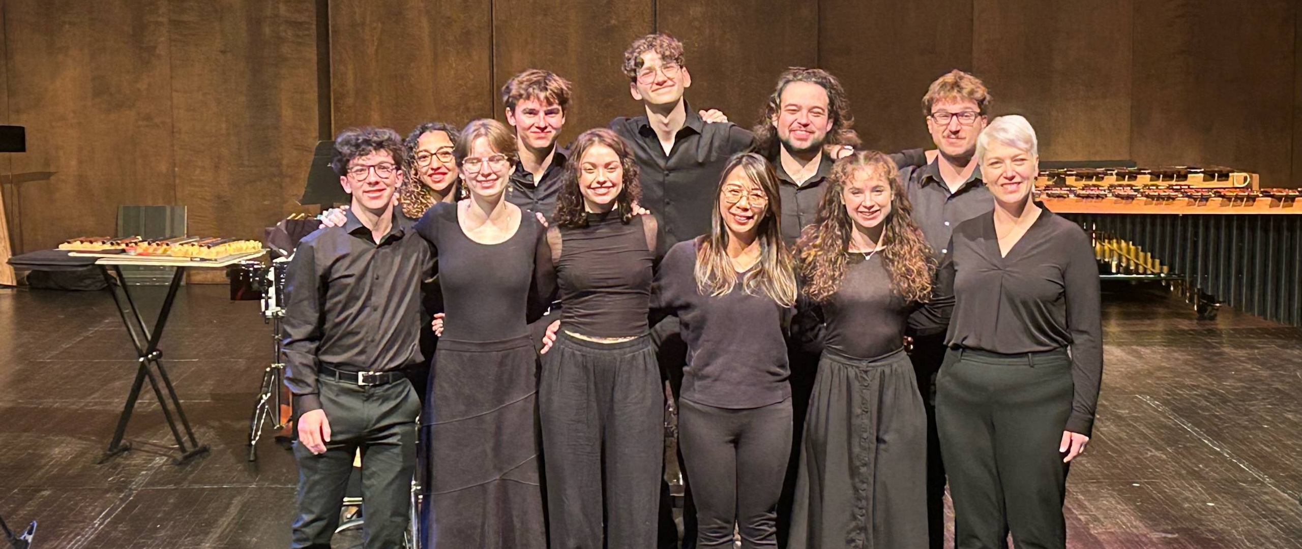 the percussion emsemble stands onstage, all dressed in black and smiling and looking at the camera.