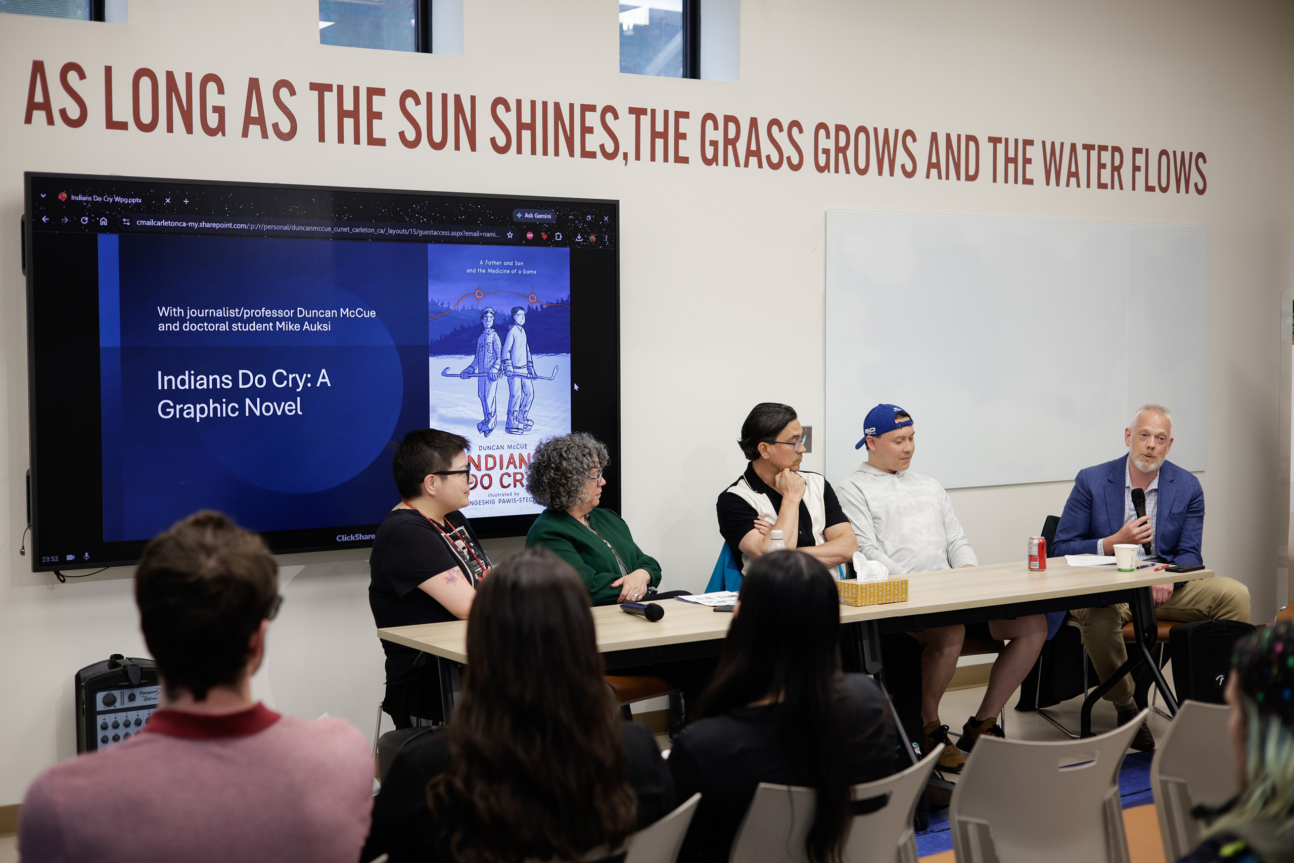a group of 5 panelists sit at a table in front of a screen while an audience looks on