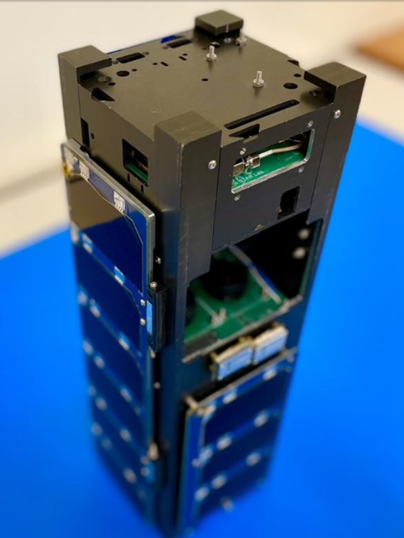 A compact, rectangular satellite device known as "CubeSat" with stacked black modules and attached solar panels, on a blue surface.