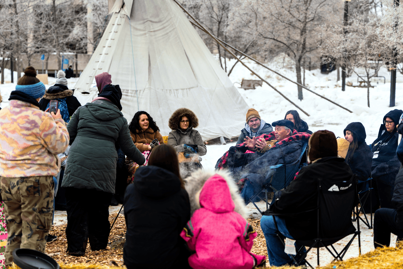 A group of people gathered around a fire outdoors in winter, seated in a circle beside a tipi.