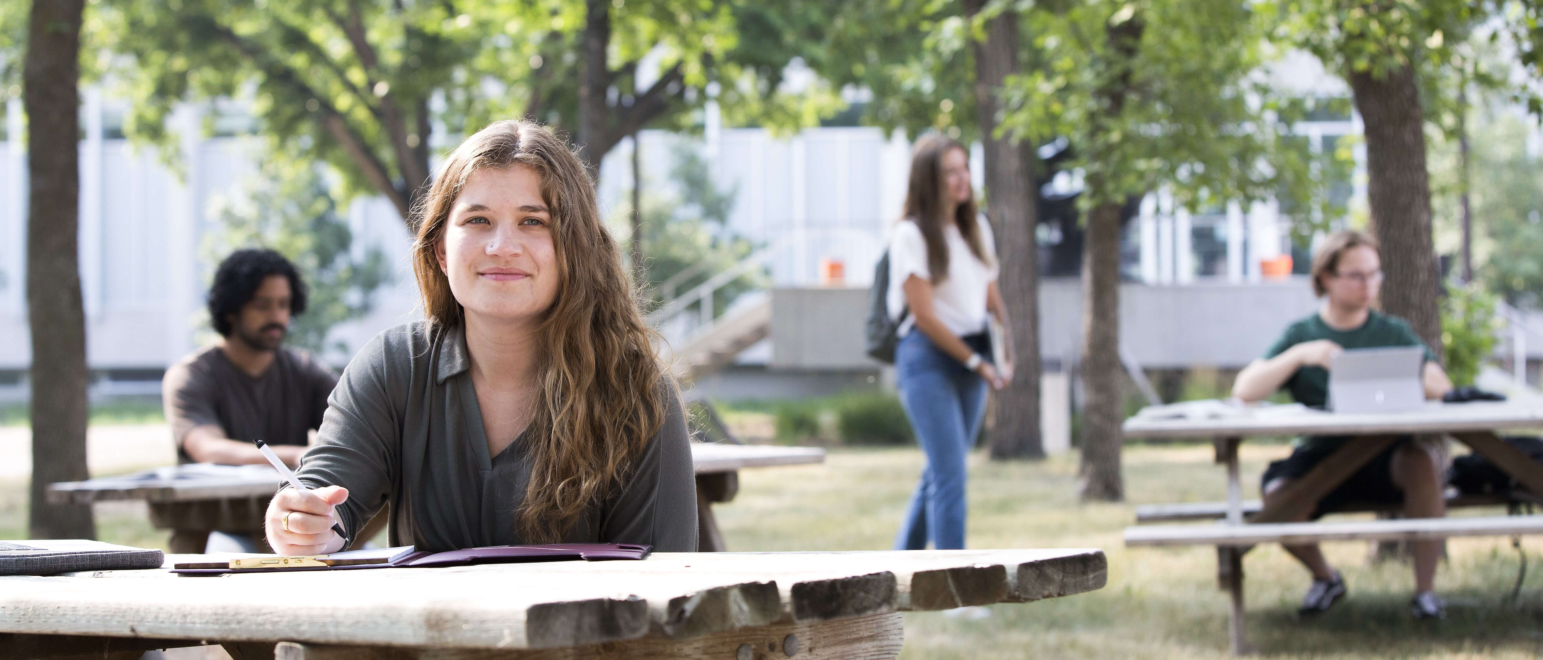 Smiling young woman sits outdoors at picnic table at Fort Garry campus.