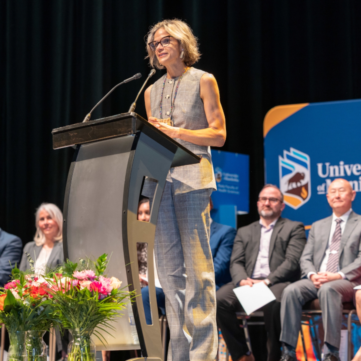 Dr. Sara Goulet speaks at a lectern. Eight health-care professionals sit in chairs behind her. 