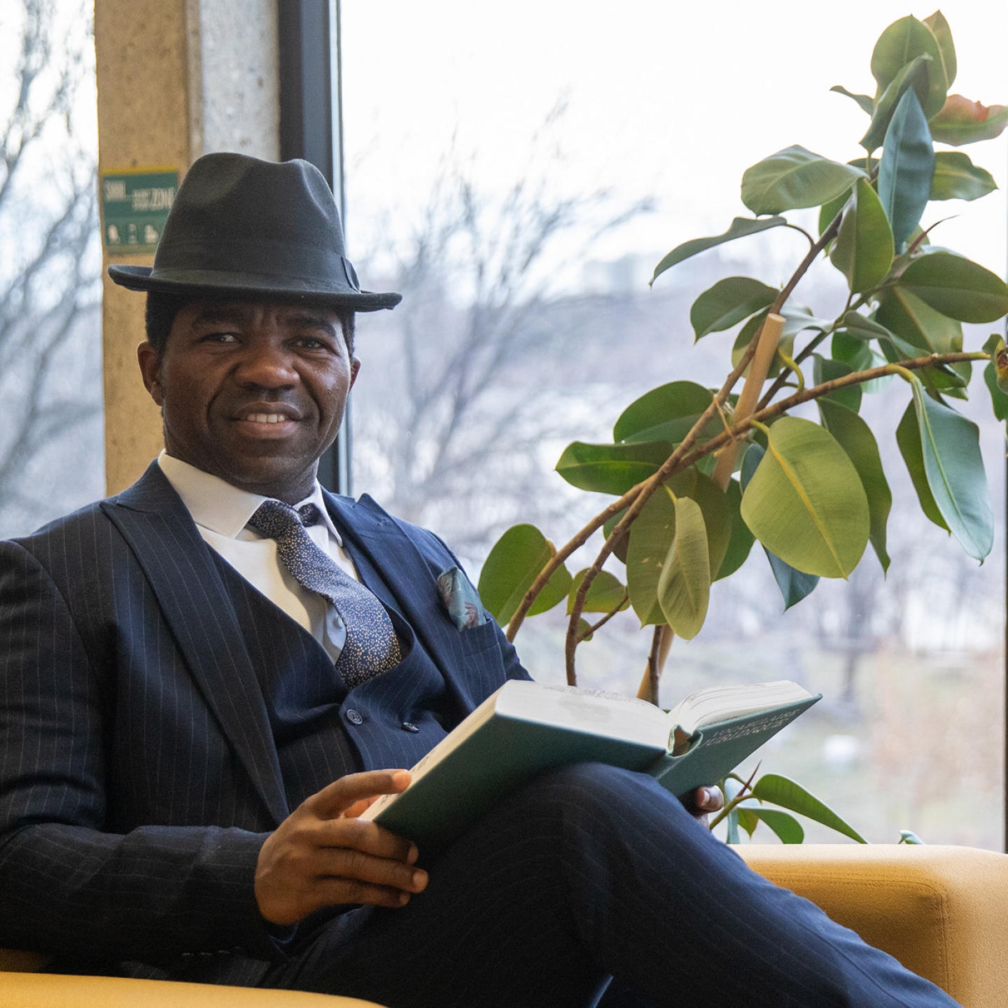 A man in a suit wears a hat and smiles at the camera holding a book, sitting in a chair in front of a large window.