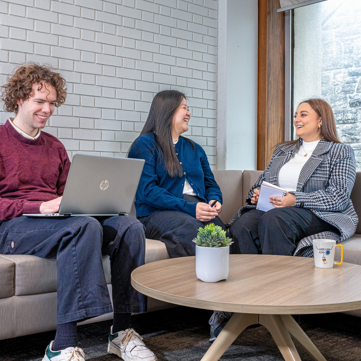 Three students sitting on a sofa in a professional environment. All wearing employee badge lanyards. One typing on a laptop. 