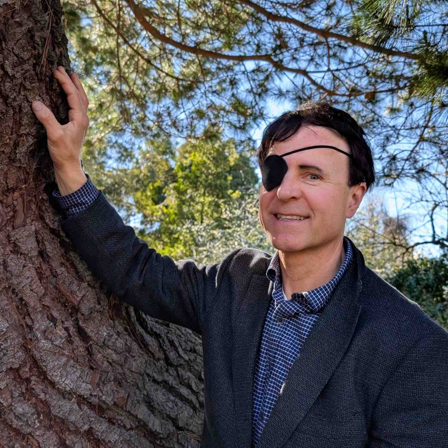 a smiling man in blue suit jacket rests an arm against a large tree