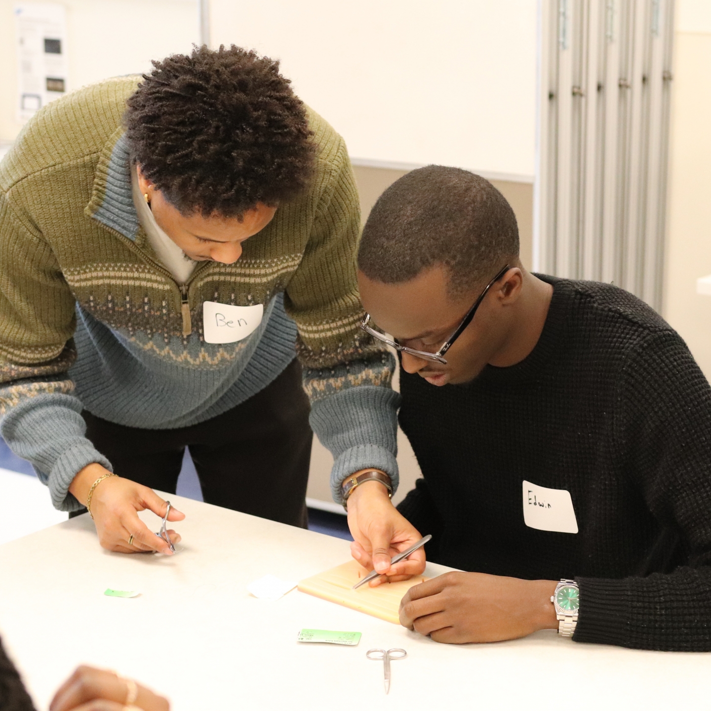 The medical student is holding scissors in one hand and tweezers in the other. He is showing the undergraduate student how to suture.