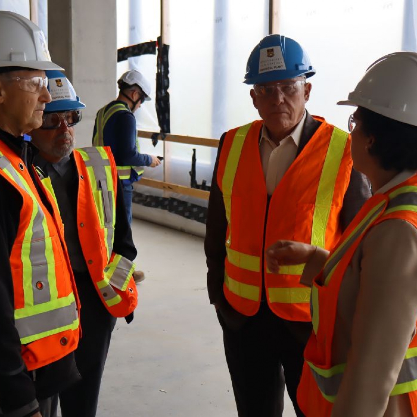 Four people wearing hard hats, safety glasses, high-vis vests talk in a construction area.