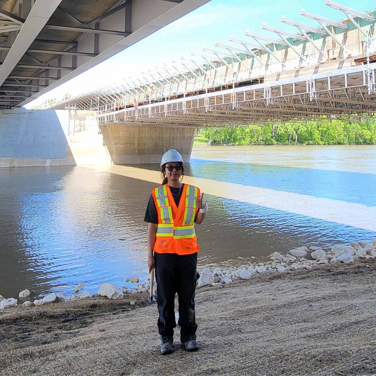 Young woman in construction gear stands under bridge with river in background.