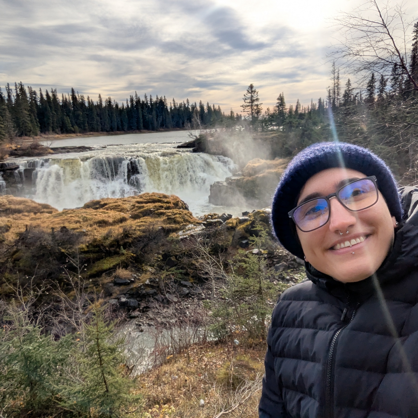 A person takes a selfie and smiles at the camera, in the background is Pisew Falls surrounded by pine trees.