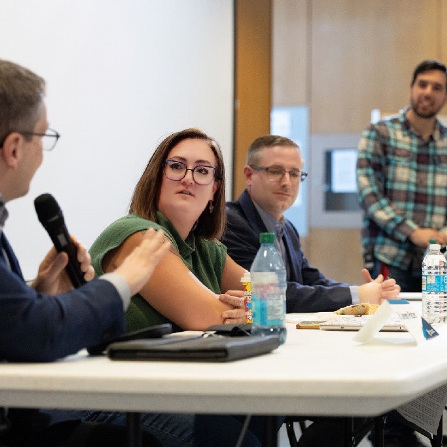 Panel discussion with three speakers seated at a table and one standing behind, as a man speaks into a microphone during an indoor event.