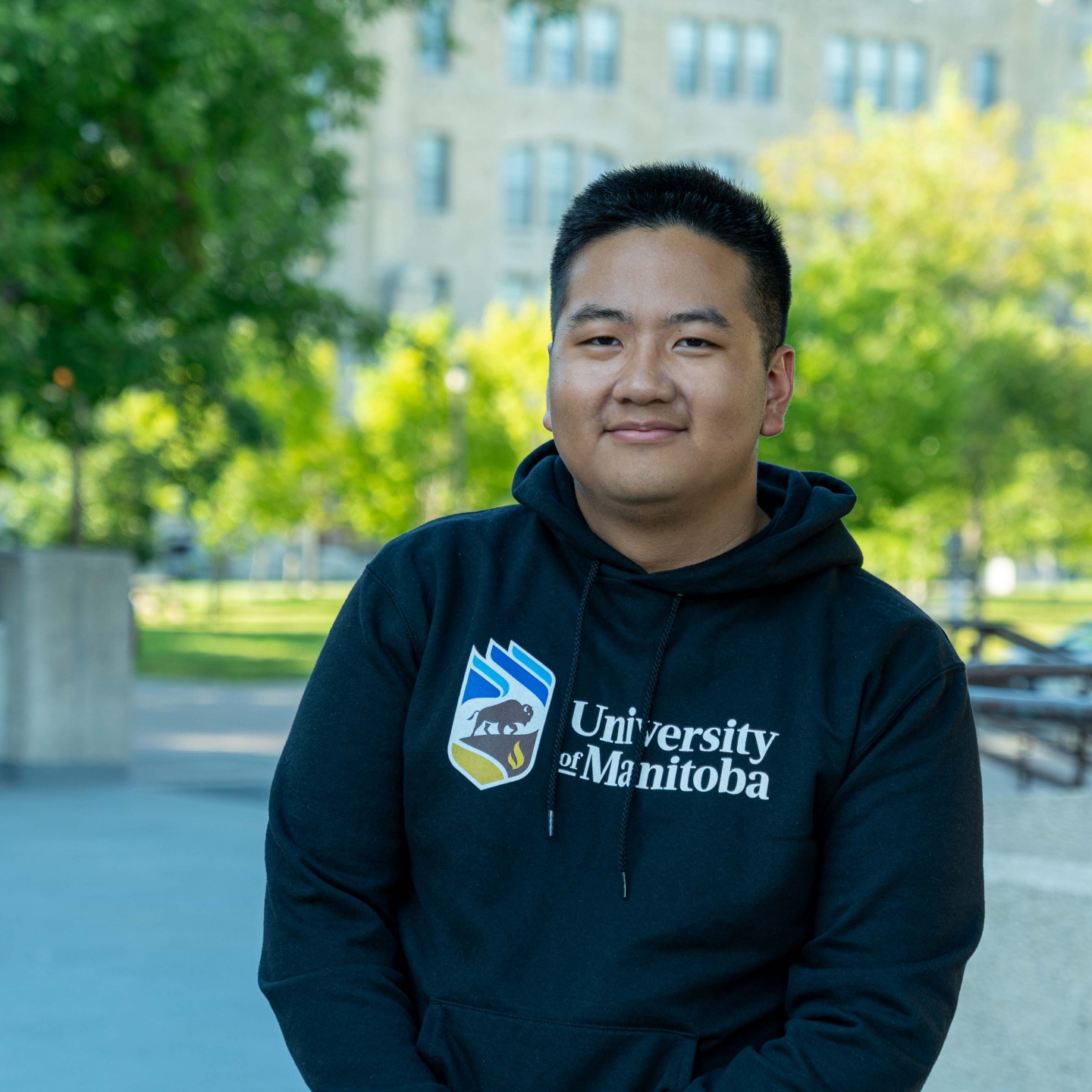 A student wearing a University of Manitoba hoodie posing in front of a background of university buildings and green trees.