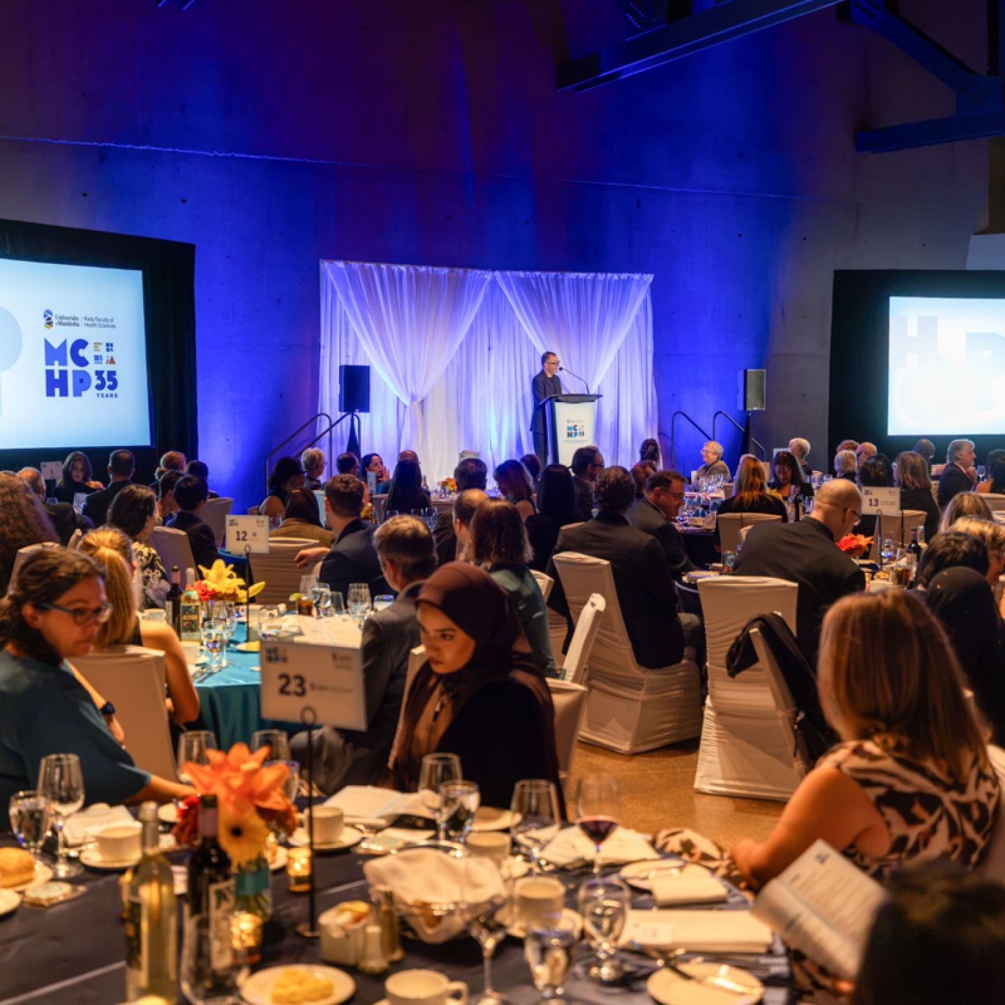Gala dinner event with a speaker at a podium. Two large screens display the text: “University of Manitoba, Rady Faculty of Health Sciences, MCHP, 35 Years.”