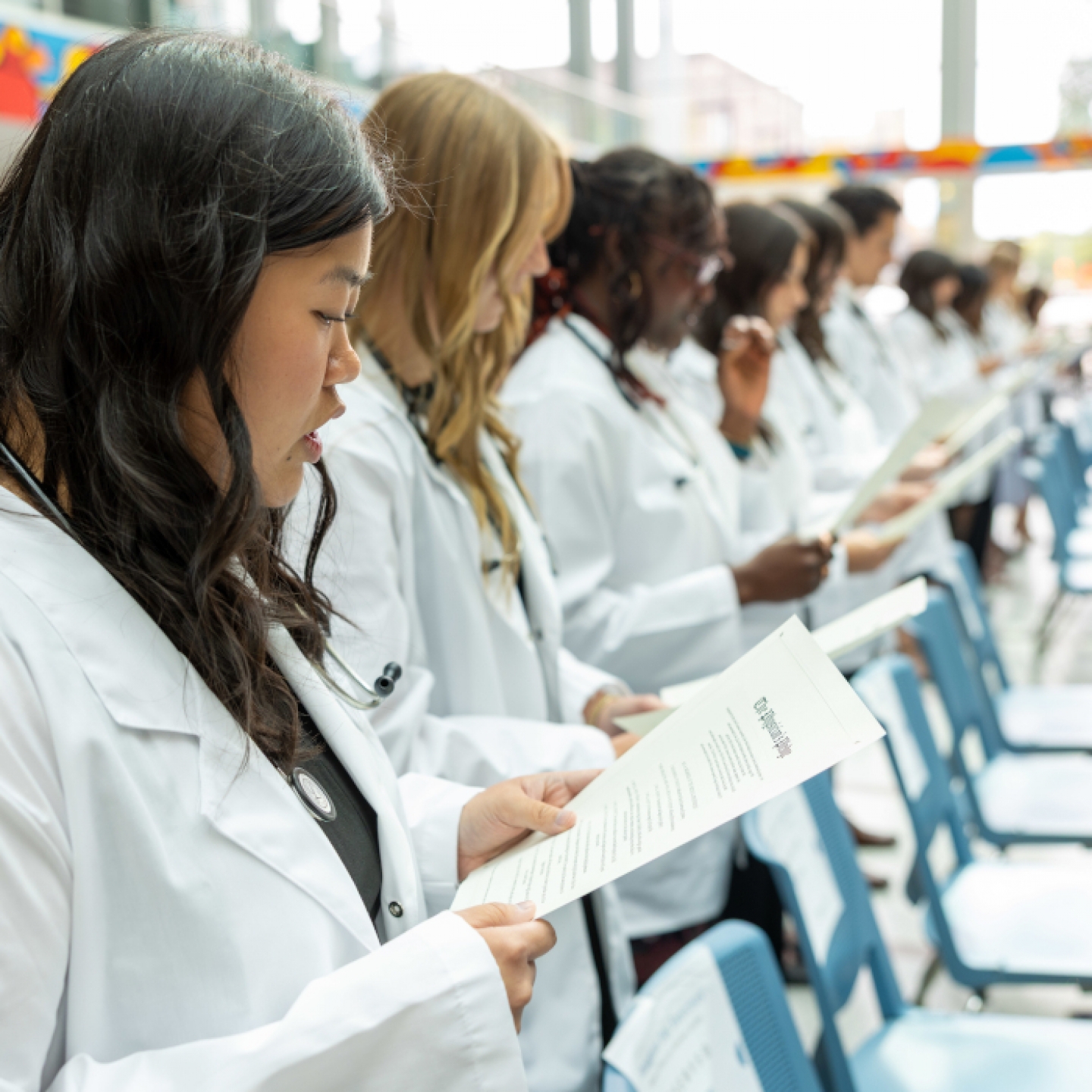 Students wearing white coats and stethoscopes around their necks, read off of sheets of paper.