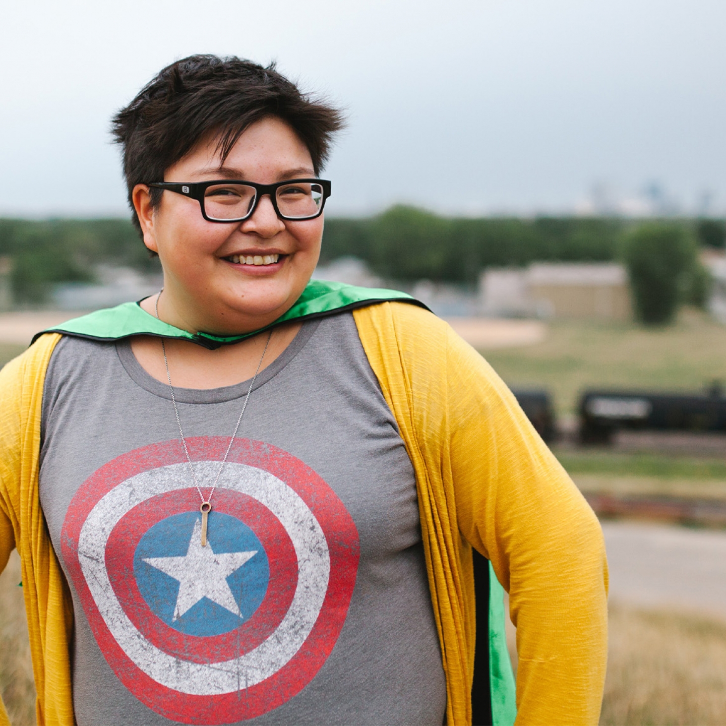 Woman wearing glasses, a tshirt with the Captain America shield on it and a cape, stands with her hands on her hips.