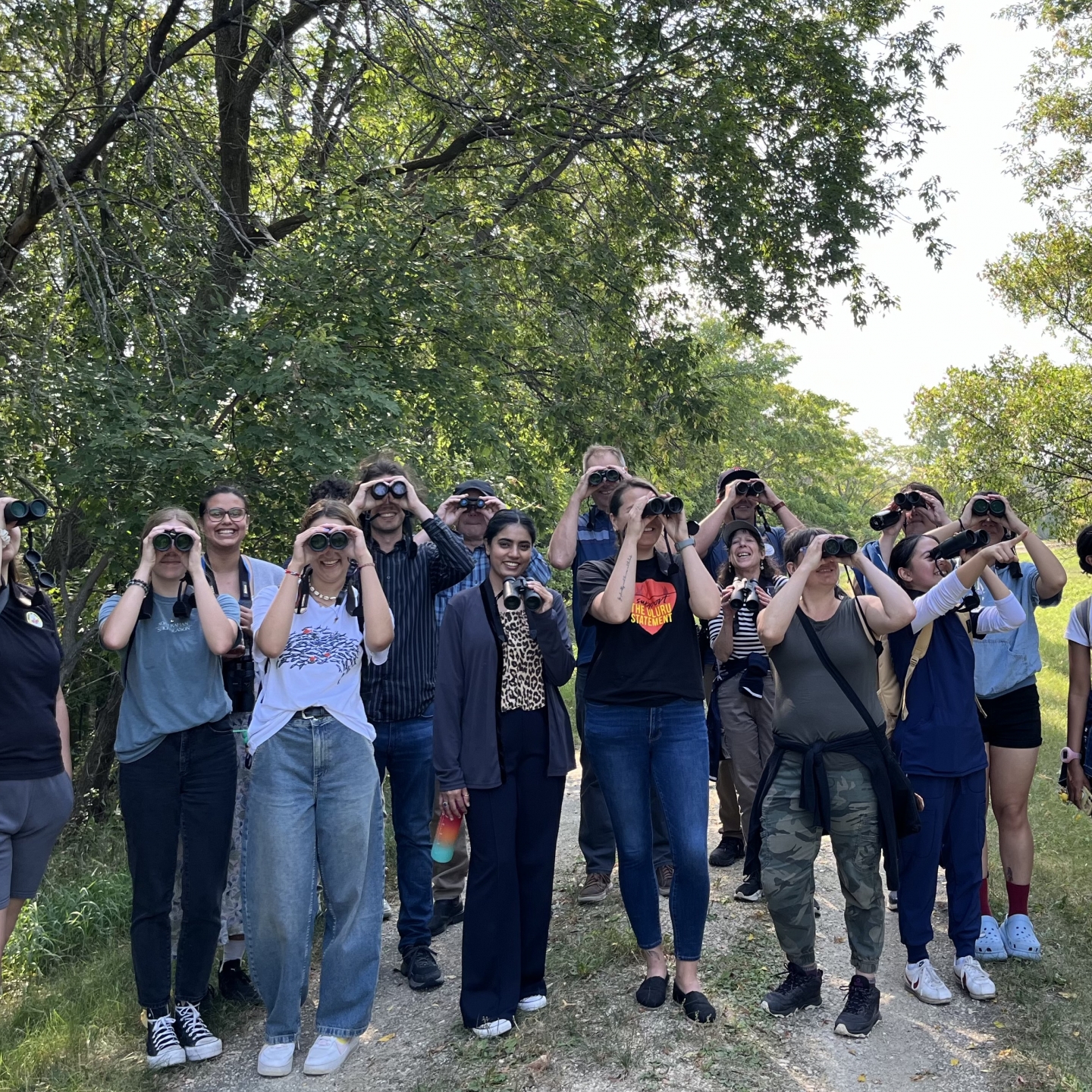 group of people holding up binoculars to their eyes and smiling, with trees in the background.