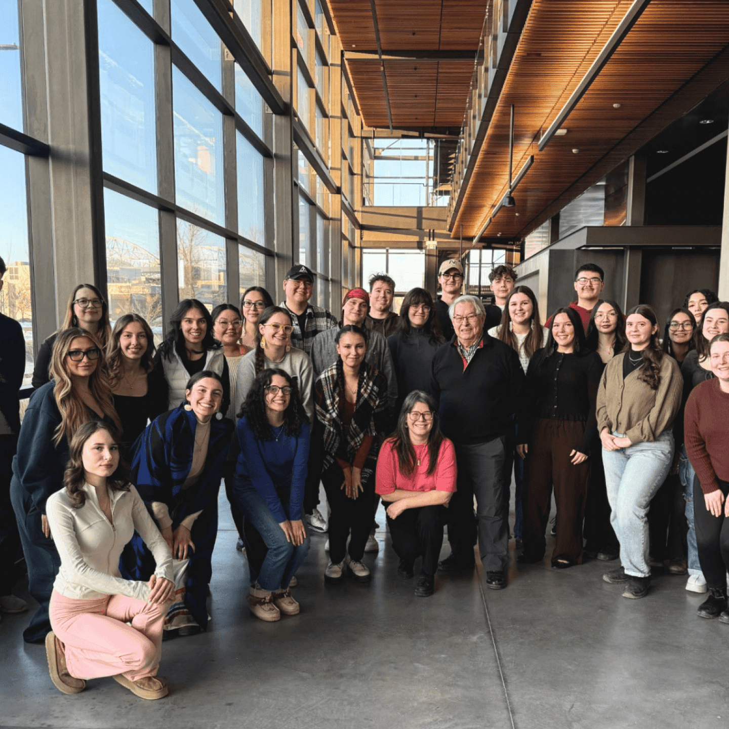 A group of Indigenous students and staff posing together in a bright indoor atrium with large windows.