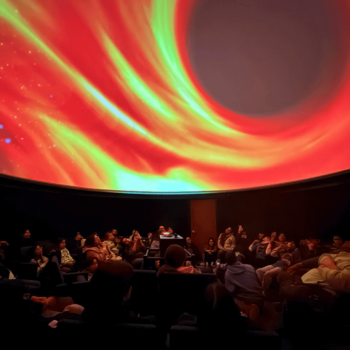 A group of attendees seated in a dome theatre, watching a vivid projection of a swirling cosmic scene resembling a black hole.