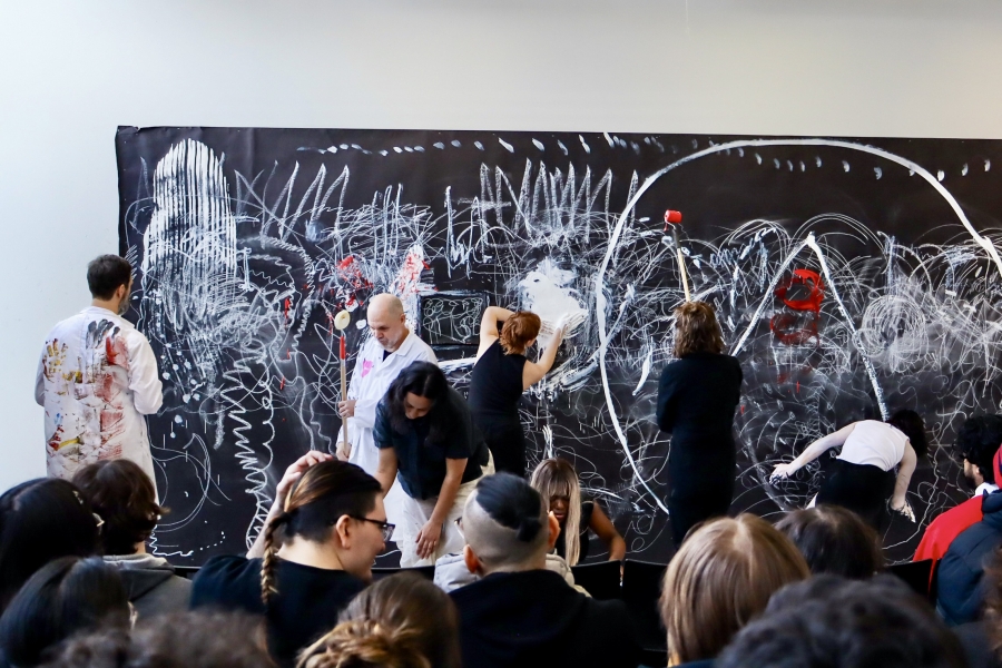 A wide view shows students painting a collaborative artwork during a live performance with an audience present.