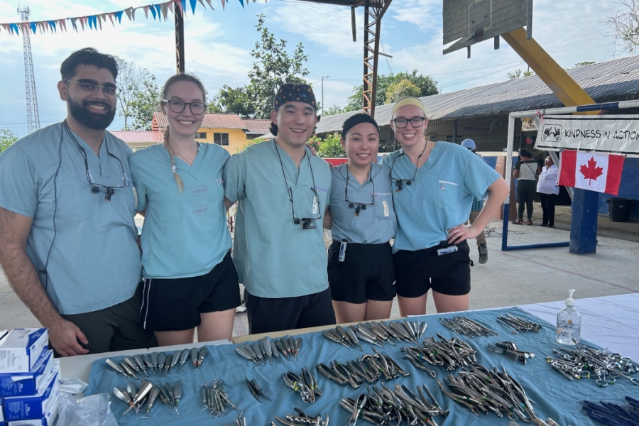 Five dental students behind a table of dental instruments. A Canadian flag and ‘Kindness in Action’ sign in the background