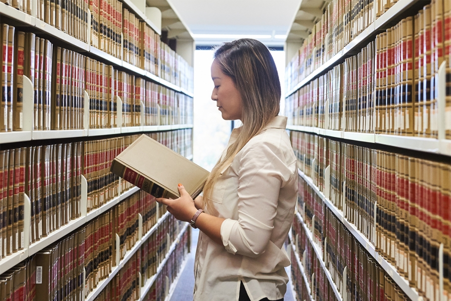 A law student holds a book in her hand as she stands in a long row of library shelves full of books.