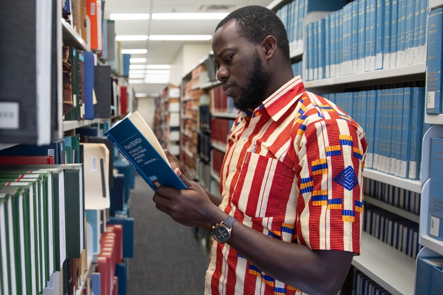 A student in a white and red shirt reads a book in front of library shelves
