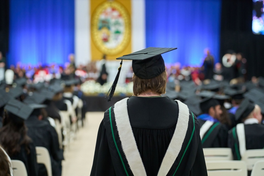 A UM grad walks down the centre aisle at Convocation. They wear a cap and gown and their back is to the camera.