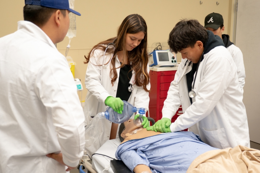 Two people wearing white coats and stethoscopes practice on a training manikin.