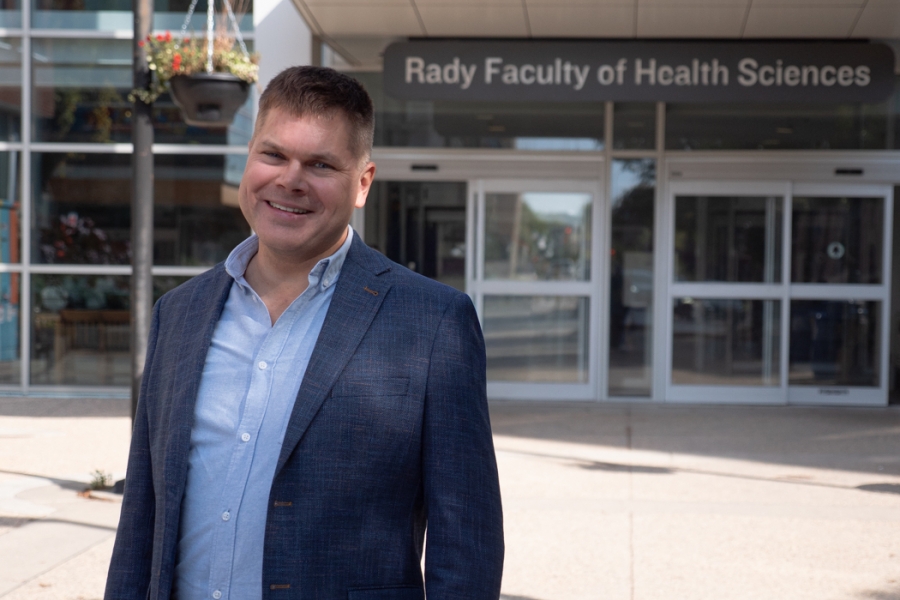 Barret Monchka smiles at the camera while standing outside the Brodie Centre. In the background, the entrance to the Rady Faculty of Health Sciences is visible.