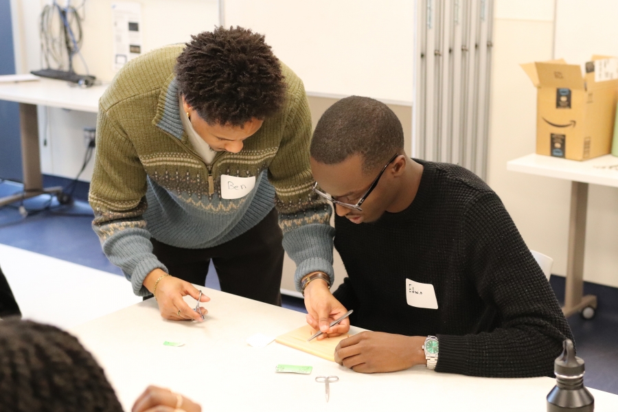 The medical student is holding scissors in one hand and tweezers in the other. He is showing the undergraduate student how to suture.