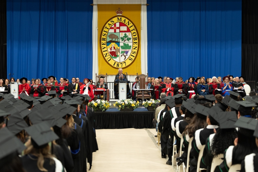Convocation ceremony seen from back of room, with rows of students seated facing colourful stage with podium.