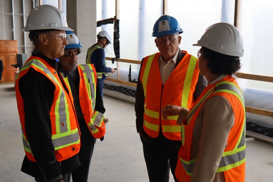 Four people wearing hard hats, safety glasses, high-vis vests talk in a construction area.
