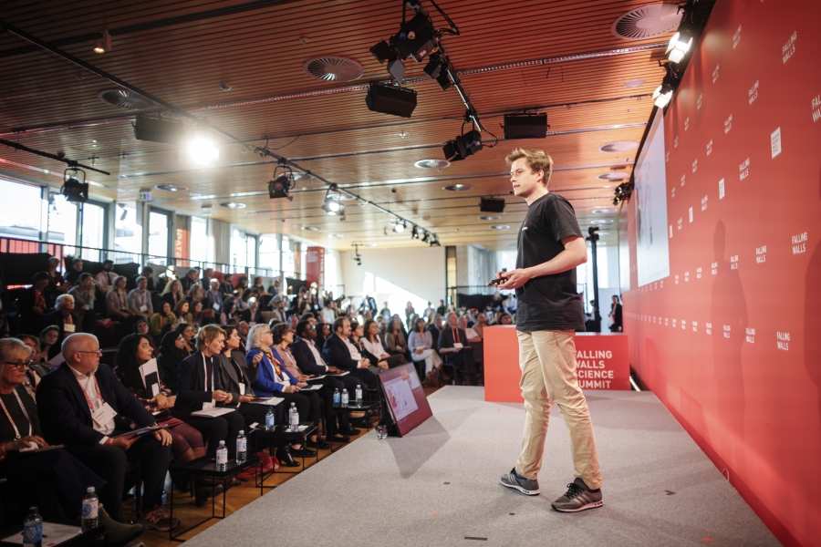 Presenter on stage at a Falling Walls Science Summit event, addressing a seated audience in a conference hall.