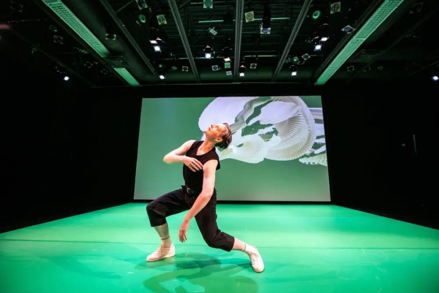 Olafson moves across a green-lit stage, their body mirrored by a projected digital figure behind them in an extended-reality performance.