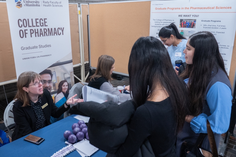 Two people sit behind a table at a College of Pharmacy booth. Three students stand in front, chatting with them.