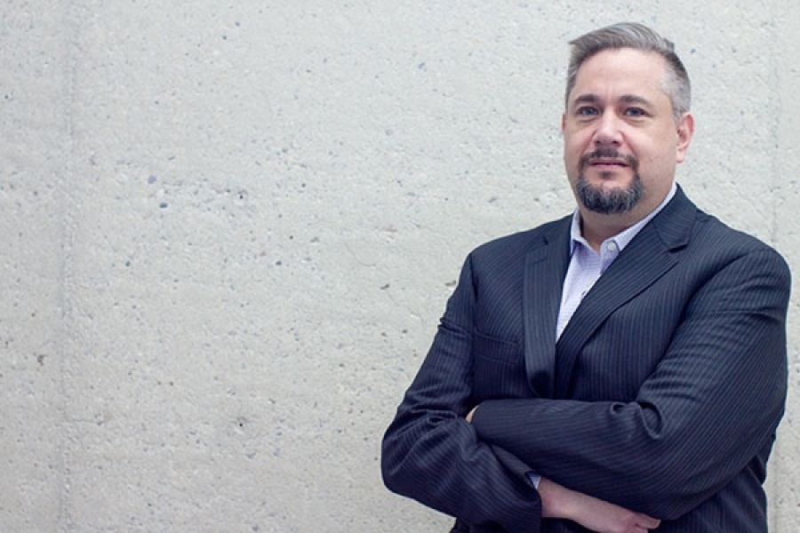 a man in a suit jacket stands in front of a concrete wall to the right of the frame.